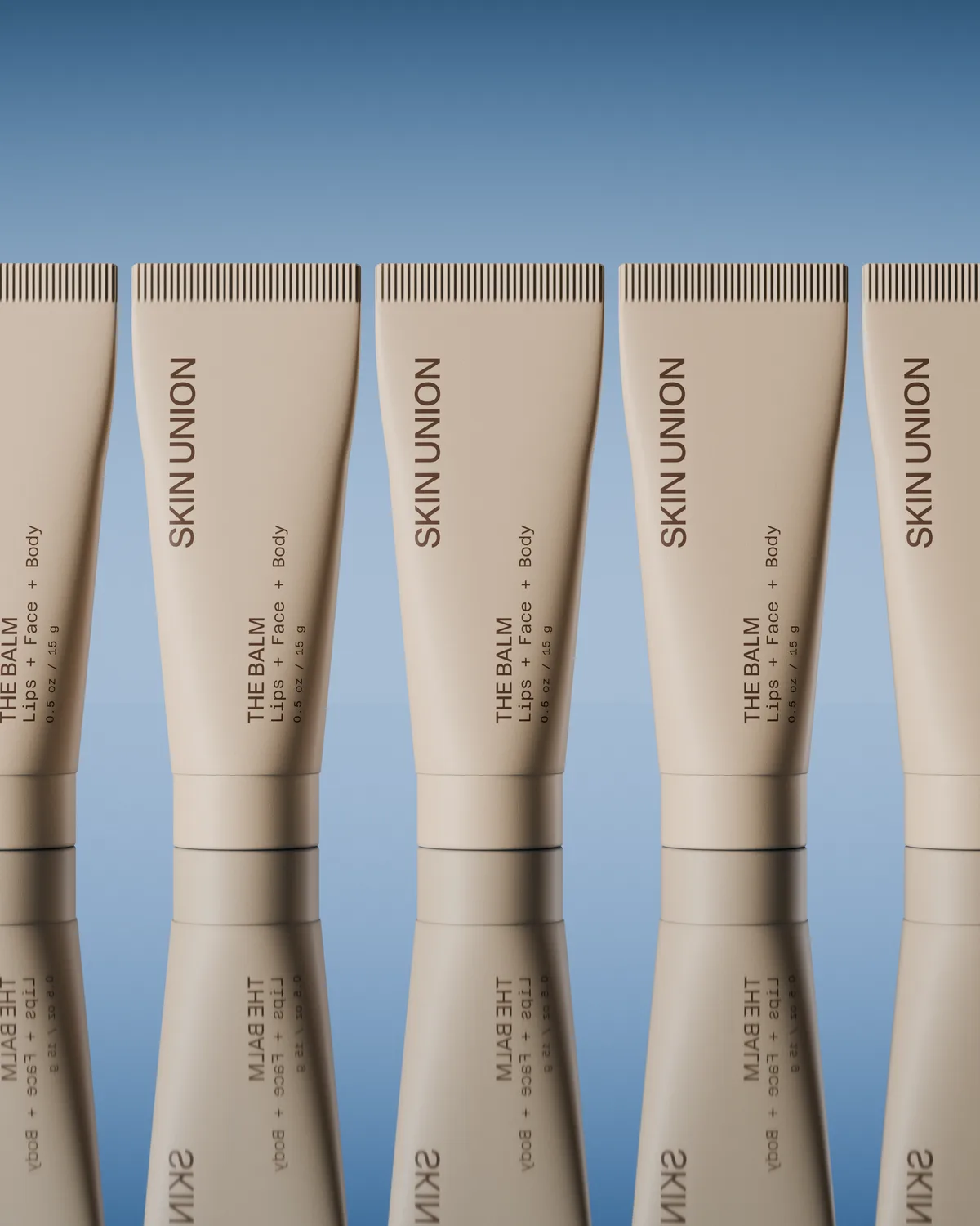 A close-set row of tubes of The Balm, reflected on a glassy surface under a soft blue sky