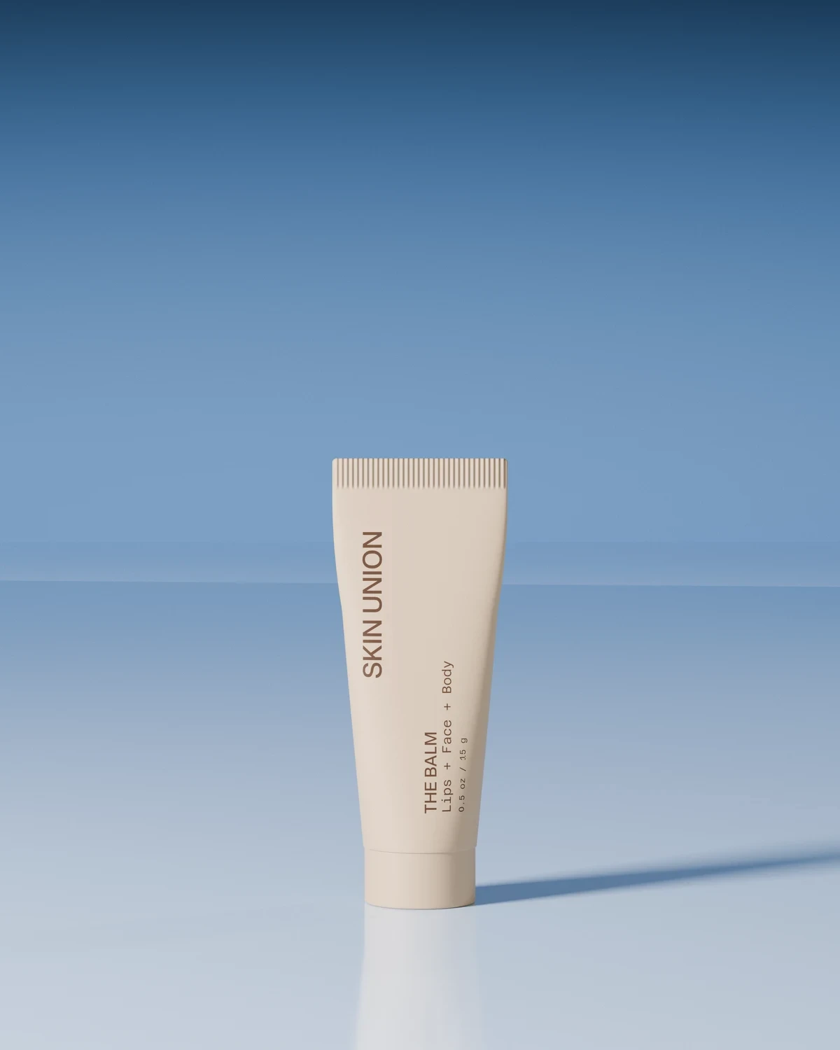 The Balm — a small pale tube labelled Lips + Face + Body, standing upright on a soft white surface against a blue backdrop